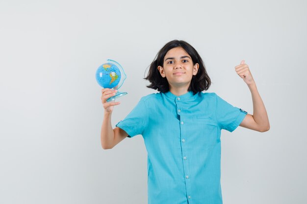 Young lady holding mini globe while showing winner gesture in blue shirt and looking satisfied.
