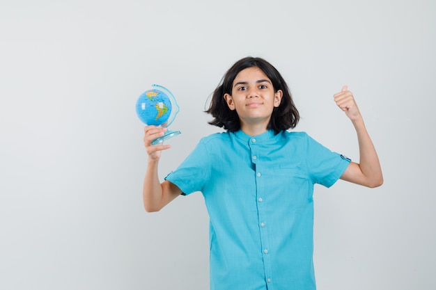 Молодая леди держит мини-глобус, показывая жест победителя в голубой рубашке и выглядит довольным.