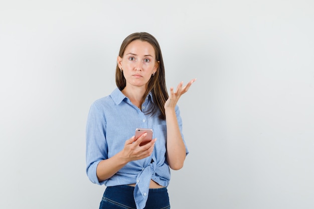 Young lady holding mobile phone in blue shirt, pants and looking confused.