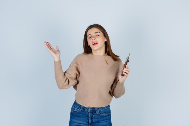 Молодая дама держит мобильный телефон в бежевом свитере, джинсах и выглядит счастливым. передний план. Бесплатные Фотографии