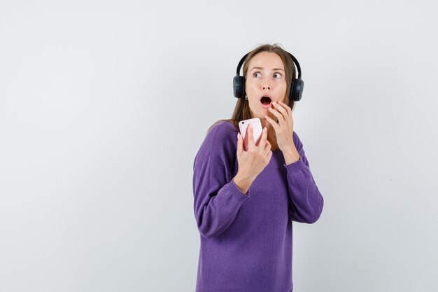 Young lady holding mobile phone in violet shirt and looking surprised , front view.