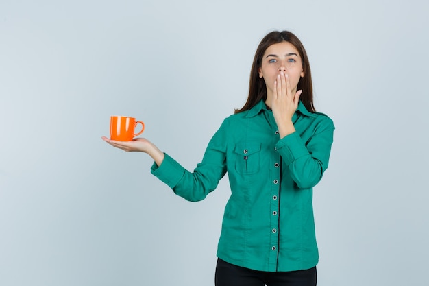 Young lady holding orange cup of tea, keeping hand on mouth in shirt and looking attractive , front view.