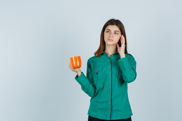 Young lady holding orange cup of tea, raising hand near face in shirt and looking pensive. front view.