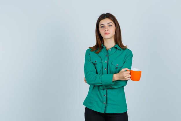 Giovane signora che tiene tazza di tè arancione in camicia e guardando fiducioso, vista frontale.