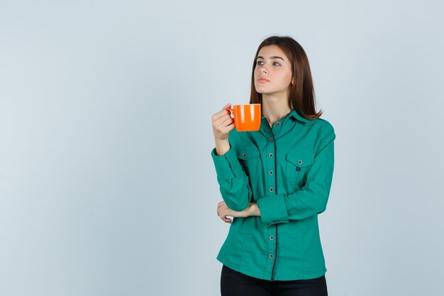 Young lady holding orange cup of tea in shirt and looking pensive , front view.