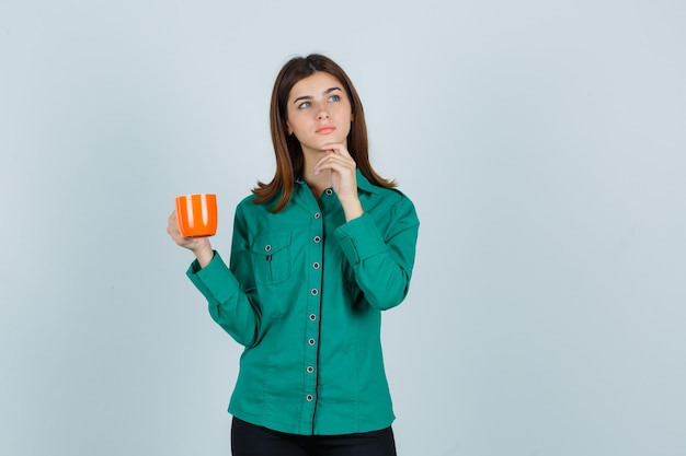 Young lady holding orange cup of tea in shirt and looking pensive , front view.