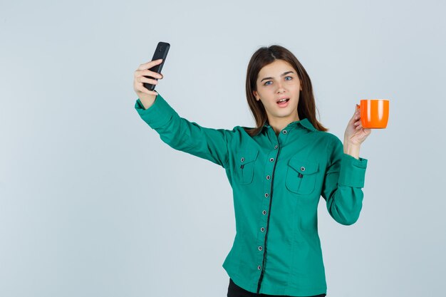 Young lady holding orange cup of tea, taking selfie with mobile phone in shirt and looking confident. front view.