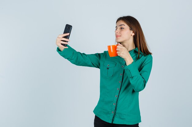 オレンジ色のお茶を持って、シャツに携帯電話で自分撮りを取り、自信を持って、正面図を見て若い女性。