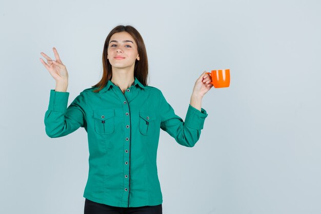 Giovane signora che tiene una tazza di tè arancione mentre mostra il numero tre in camicia e sembra soddisfatto, vista frontale.