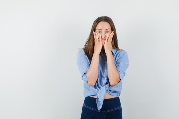 Young lady holding palms on cheeks in blue shirt, pants and looking helpless