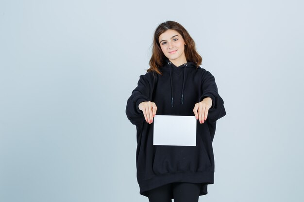 Young lady holding paper in front of her in oversized hoodie, pants and looking elegant. front view.