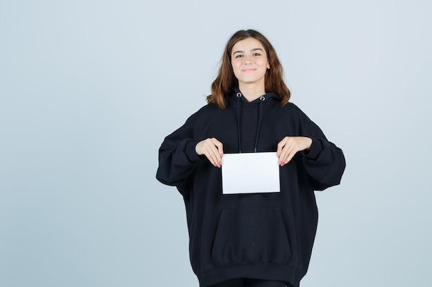 Young lady holding paper in front of her in oversized hoodie, pants and looking happy , front view.