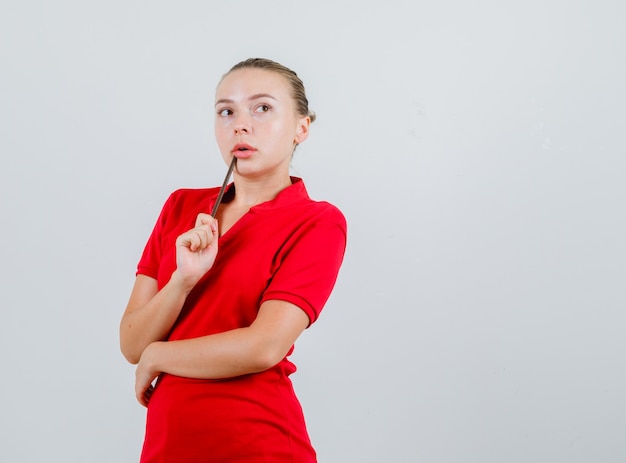Молодая леди держит карандаш возле рта в красной футболке и выглядит задумчиво