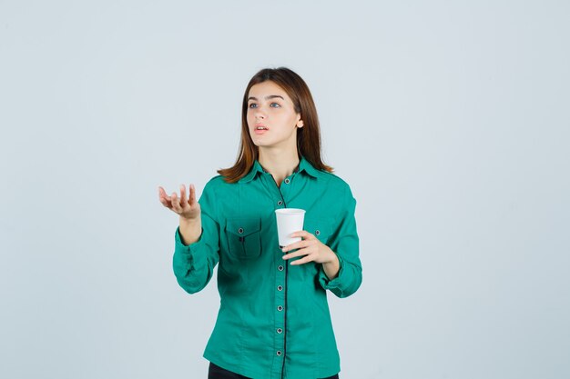 Молодая леди держит пластиковую чашку кофе, вопросительно протягивает руку в рубашке и выглядит сосредоточенной. передний план.