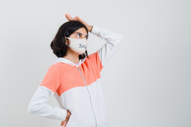 Young lady in hoodie, face mask putting hand on her head and looking tired