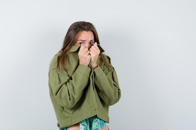 襟の後ろに顔を隠し、恐怖を探している緑のジャケットの若い女性、正面図。 無料写真