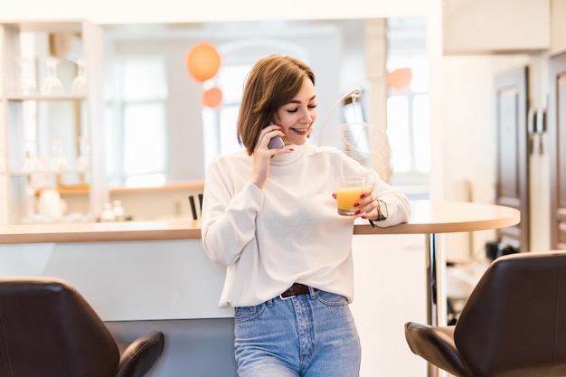 Young lady is standing near bar chair in the kitchen talking on the phone and holding a glass with orange juice