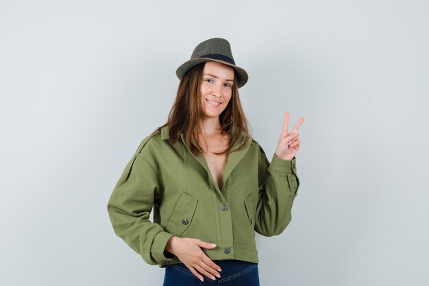 Young lady in jacket pants hat showing v-sign and looking confident  