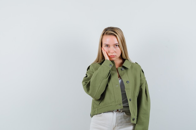 Young lady in jacket, pants holding hand on cheek and looking sad , front view.