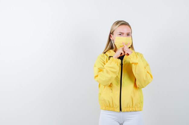 Young lady in jacket, pants, mask showing silence gesture with crossed fingers forming an x and looking careful , front view.