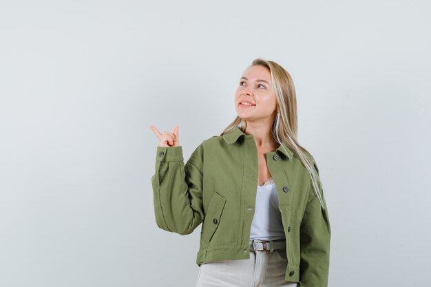 Young lady in jacket, pants pointing at upper left corner and looking happy , front view.