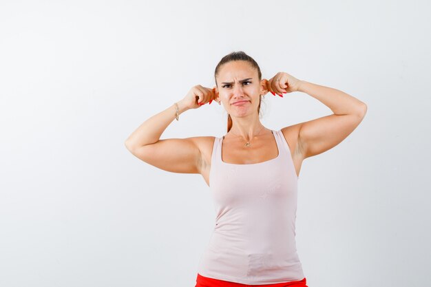 Young lady keeping ears with hands in beige tank top and looking funny. front view.