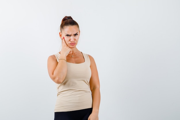 Young lady keeping finger on cheek, curving lips, frowning face in beige tank top and looking pensive , front view.