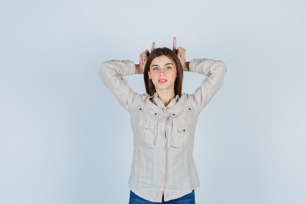 Young lady keeping fingers as bull horns in casual, jeans and looking funny. front view.