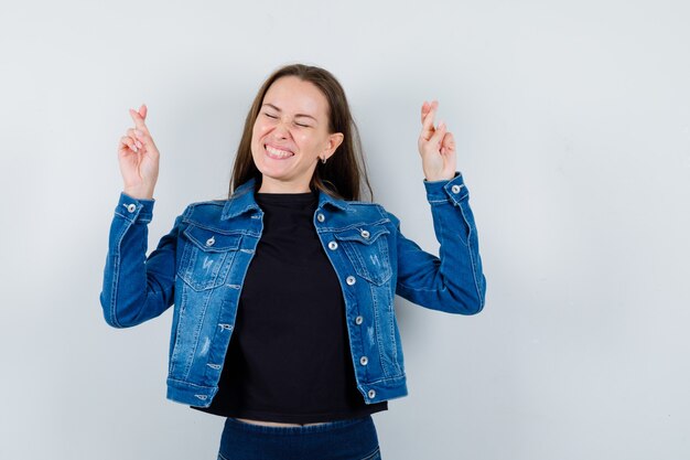 Young lady keeping fingers crossed in blouse, jacket and looking happy. front view.