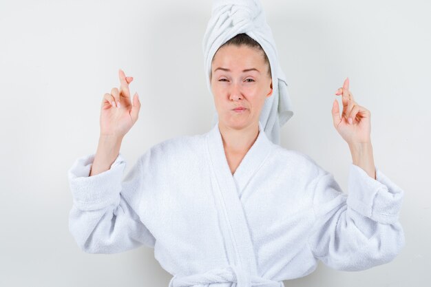 Young lady keeping fingers crossed in white bathrobe, towel and looking dissatisfied , front view.