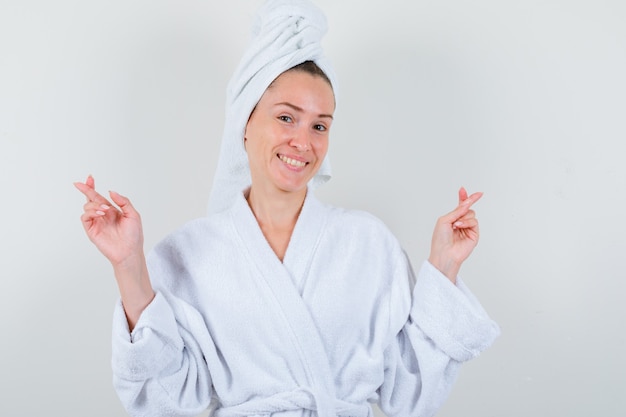 Young lady keeping fingers crossed in white bathrobe, towel and looking joyful. front view.