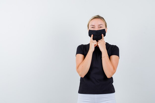 Young lady keeping fingers on her medical mask in black t-shirt and looking cheerful , front view.