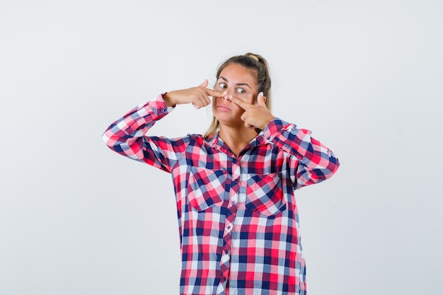 Young lady keeping fingers on nose in checked shirt and looking pensive. front view.