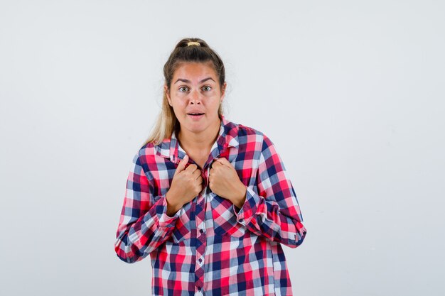 Young lady keeping fists on chest in checked shirt and looking puzzled , front view.