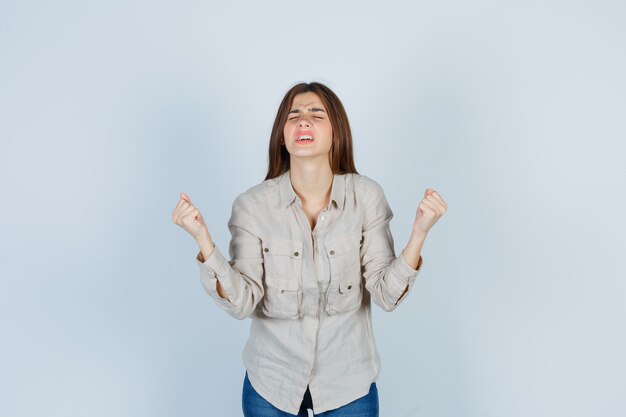 Young lady keeping fists clenched, closing eyes in casual, jeans and looking frustrated. front view.