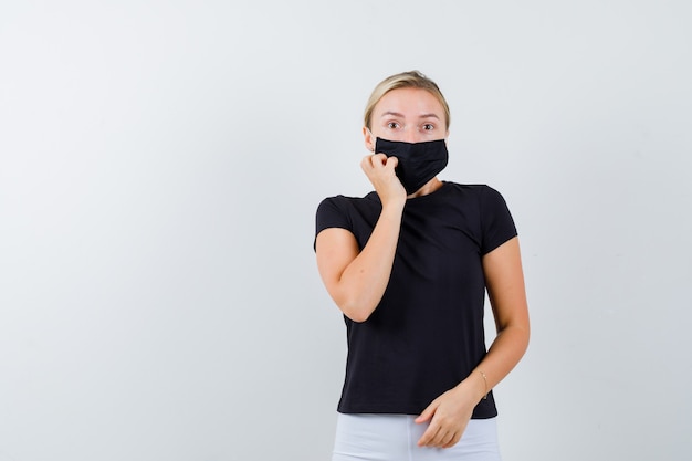 Young lady keeping hand on cheek in black t-shirt, mask and looking astonished , front view.
