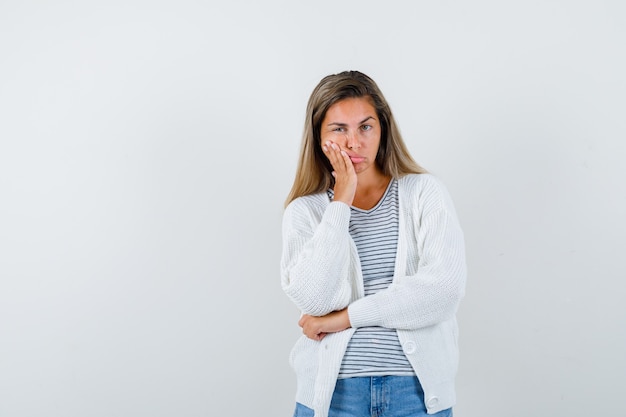 Young lady keeping hand on cheek in t-shirt, jacket, jeans and looking downcast , front view.