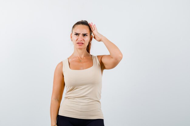 Young lady keeping hand on head in beige tank top and looking pensive. front view.