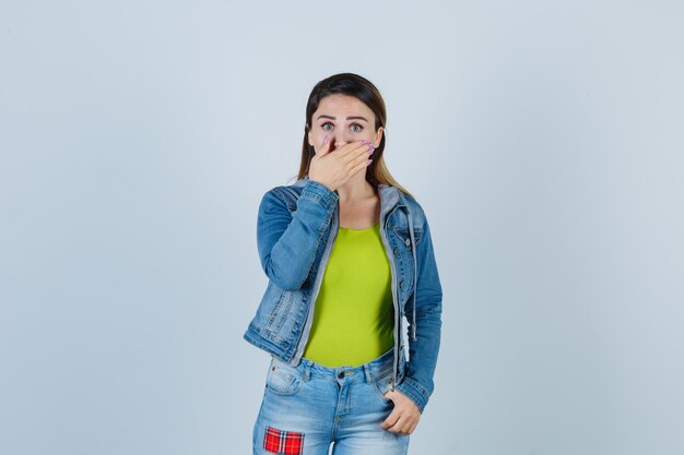 Young lady keeping hand on mouth in denim outfit and looking scared , front view.