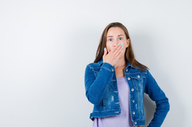 Young lady keeping hand on mouth in t-shirt, jacket and looking surprised , front view.