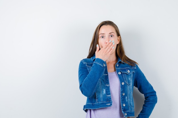 Young lady keeping hand on mouth in t-shirt, jacket and looking surprised , front view.