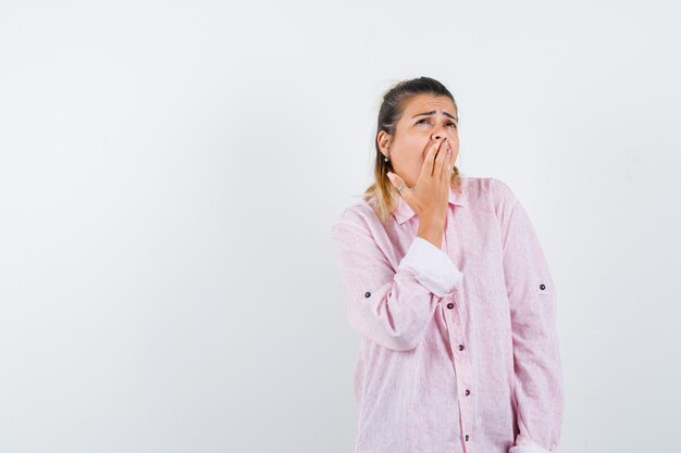 Young lady keeping hand on mouth while yawning in pink shirt and looking sleepy