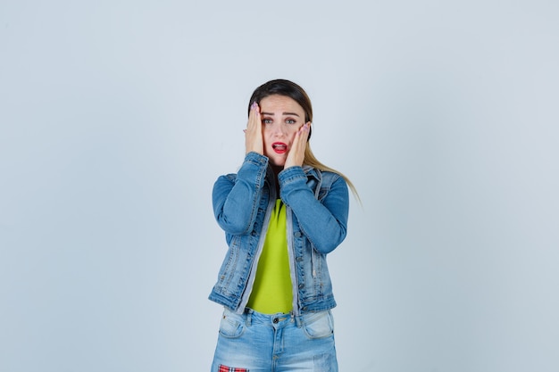 Young lady keeping hands on cheeks in denim outfit and looking anxious. front view.