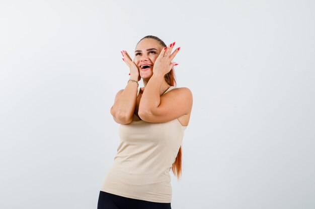 Young lady keeping hands on cheeks in tank top and looking happy , front view.