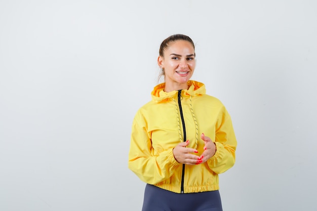 Young lady keeping hands in front of her in yellow jacket and looking cheerful. front view.