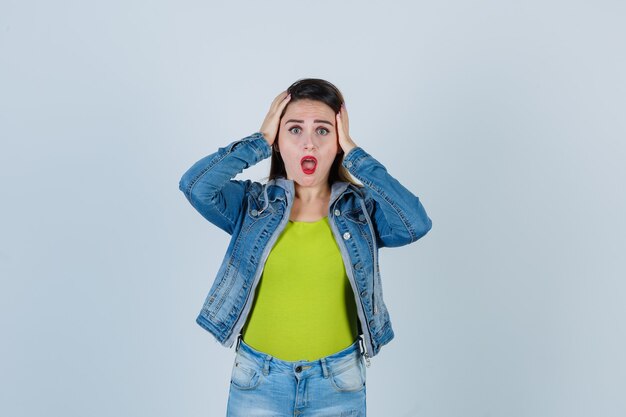 Young lady keeping hands on head in denim outfit and looking alarmed. front view.