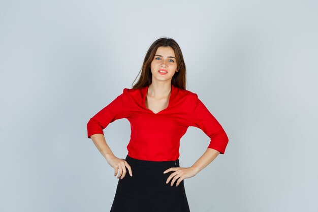 Young lady keeping hands on hip in red blouse, skirt and looking gorgeous