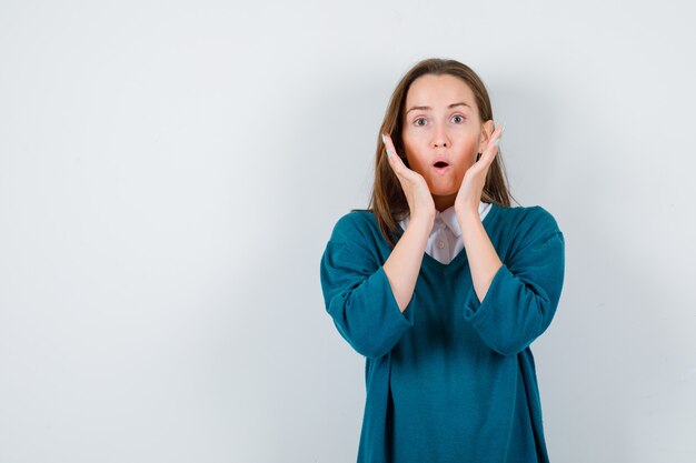 Young lady keeping hands near face in white shirt, sweater and looking wondered. front view.