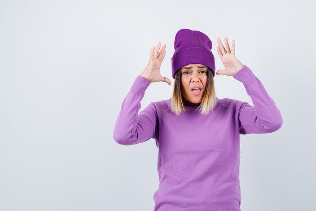 Молодая леди, держащая руки в жесте капитуляции, в фиолетовом свитере, шапочке и тревожно выглядит, вид спереди.