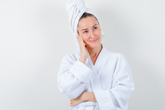 Young lady leaning cheek on hand in white bathrobe, towel and looking peaceful , front view.